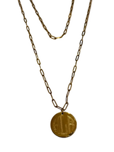 A gold-toned layered necklace featuring a paperclip chain and a circular pendant engraved with the initials 'HHA', displayed on a light marble surface.